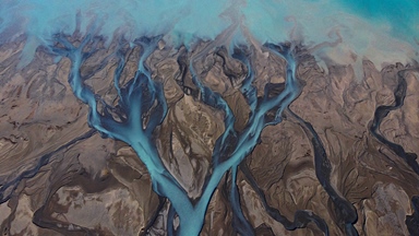 Aerial view of a braided river system with multiple bright turquoise channels flowing across a broad expanse of dark grey and brown sediment. The branching waterways weave and reconnect across the landscape, creating intricate patterns that resemble veins or tree roots when seen from above. Contrasting colours between the milky blue water and the exposed riverbed highlight the natural formations and textures of the terrain.
