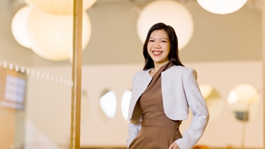 Professional portrait of a person standing indoors beside a glass partition in a modern office environment. The person is wearing a brown outfit with a light-coloured cropped jacket and is positioned slightly off-centre with hands in pockets. Large circular pendant lights appear softly out of focus in the background, creating a warm, contemporary setting. The image uses shallow depth of field and soft lighting, drawing attention to the subject while showcasing the stylish interior design.