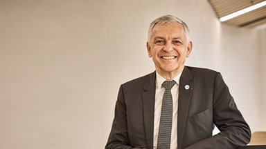 Older man in a business suit smiling at camera with one arm resting on table
