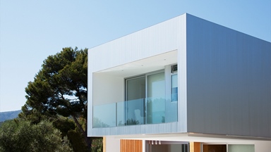 Contemporary modular house with metal cladding, expansive glazing and a first-floor balcony, positioned within a landscaped setting with mature trees.