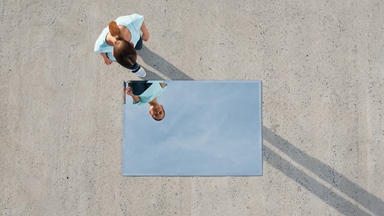 Overhead view of a person standing on a large concrete surface and looking down at a rectangular mirror placed on the ground. The mirror reflects the person's face and upper body, creating a contrast between the top-down perspective and the reflected view. Long shadows stretch across the concrete, while the minimalist composition and open space draw attention to the relationship between the person and their reflection. The image symbolises self-reflection, identity, perspective or personal growth.