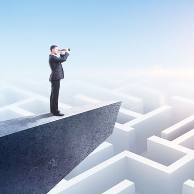 Person in business attire standing on a concrete platform above a maze, using binoculars to look ahead across the complex pathways.