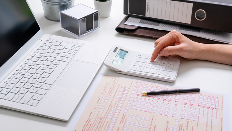 A person uses a calculator beside a laptop while completing a printed tax form on a desk. Office items, including folders, a pen and desk accessories, are arranged nearby, illustrating personal tax or financial administration.