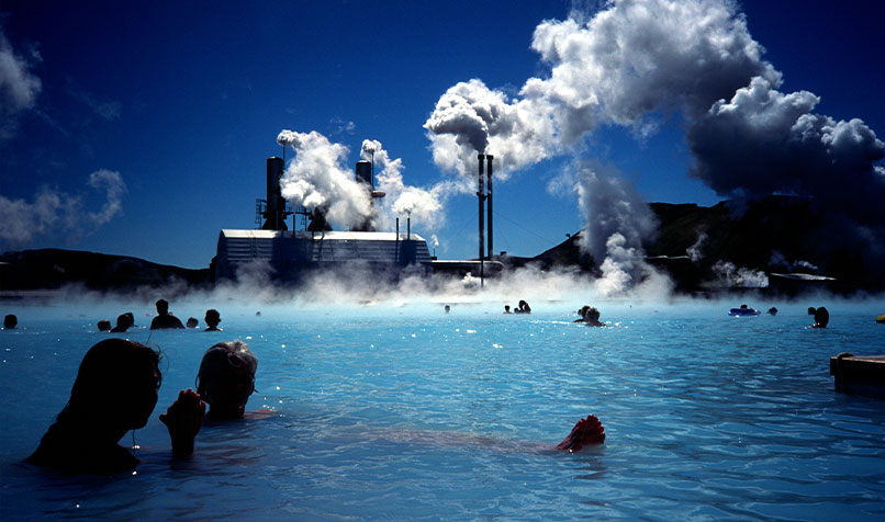 People bathing in blue lagoon with power station in background with steam coming out of chimneys
