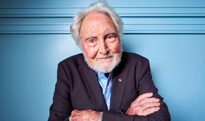 Older man with white hair and beard in a dark suit standing in front of a blue wall with arms crossed