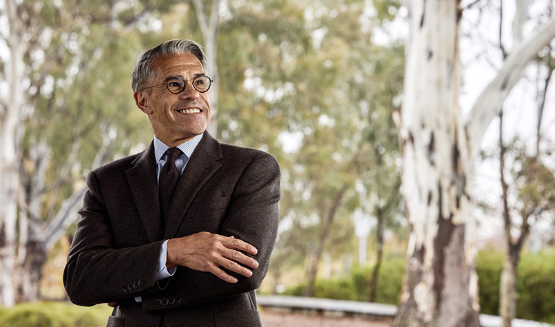 Person wearing a brown suit, dress shirt, and tie stands outdoors with arms crossed. The person is wearing round glasses and a wristwatch, and is positioned in front of a blurred background of tall trees and greenery, with soft natural light filtering through the scene.
