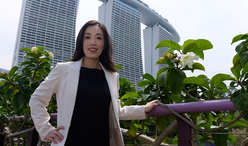 Person standing outdoors beside leafy green plants, wearing a light-colored blazer over dark clothing, with one hand resting on a railing. In the background, three tall modern buildings with a distinctive rooftop structure are visible against a bright sky.