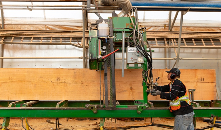 Worker wearing hearing protection, gloves and high-visibility clothing operating a timber processing machine in a wood manufacturing facility.