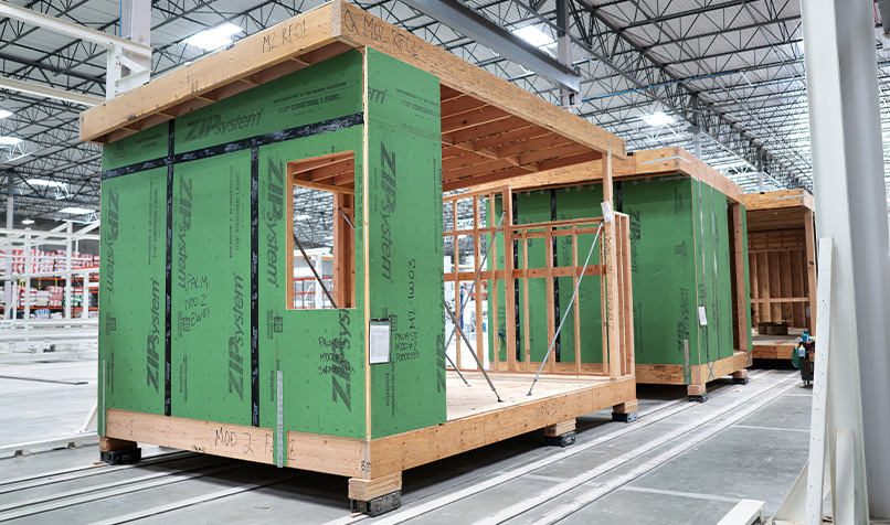 Modular home units under construction within a factory, with exposed timber frames, green wall sheathing and production equipment visible throughout the facility.