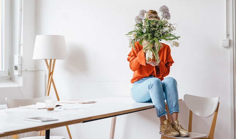 Person wearing an orange jumper and jeans, seated on a table while holding a large bouquet of flowers in front of the face, in a light and modern room.