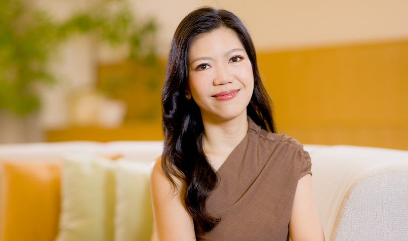 Professional portrait of a person seated on a light-coloured sofa in a softly lit indoor setting. The person has long dark hair and is wearing a sleeveless brown top. The background is intentionally blurred, featuring neutral-toned furnishings and a green plant, creating a warm and contemporary office or lounge environment. The subject is positioned centrally in the frame, with soft lighting and natural tones contributing to a polished, professional image.
