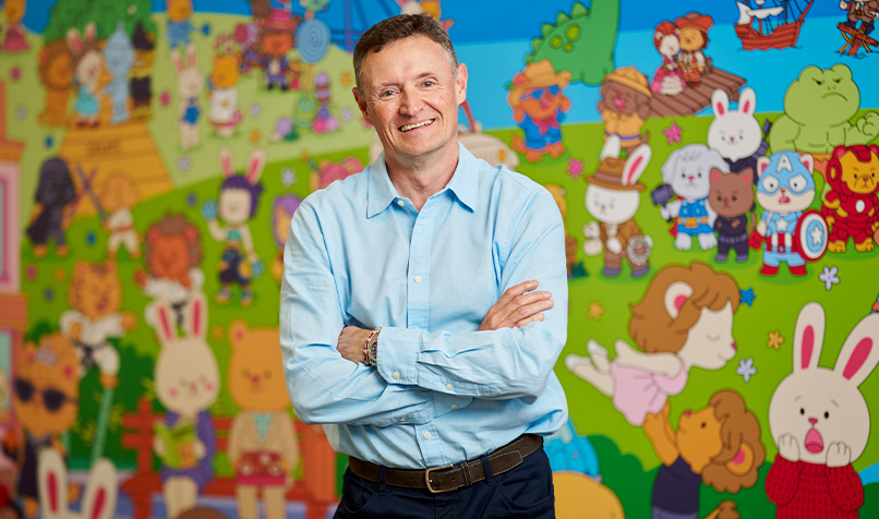 A person wearing a light blue button-down shirt stands with folded arms in front of a colourful mural featuring cartoon-style characters. The illustrated backdrop includes rabbits, bears, lions, fairy-tale figures, superheroes, animals and whimsical landscape scenes in bright shades of green, blue, pink and yellow. The person is positioned in the centre of the image, with the vibrant artwork filling the entire background and creating a playful, child-focused setting.
