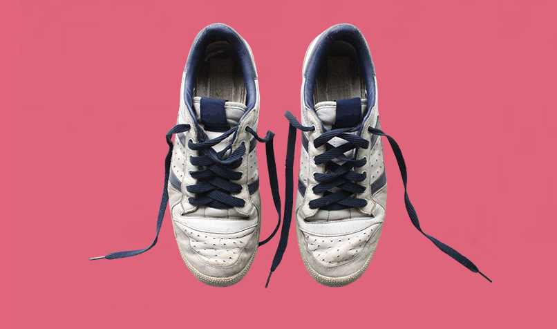 A pair of well-worn white trainers with navy blue laces is displayed against a solid pink background. The shoes are positioned side by side and viewed from above, with the untied laces extending outward on either side. Visible creases, scuff marks and signs of use on the leather surface and soles give the trainers a lived-in appearance, while the contrasting pink backdrop creates a bold, minimalist composition.