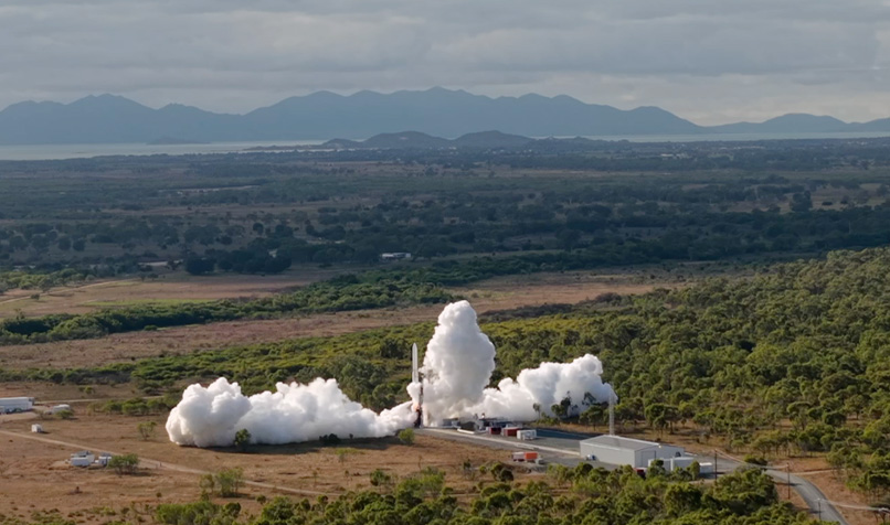Rocket launching from a ground facility, producing a large plume of white smoke across the surrounding landscape.