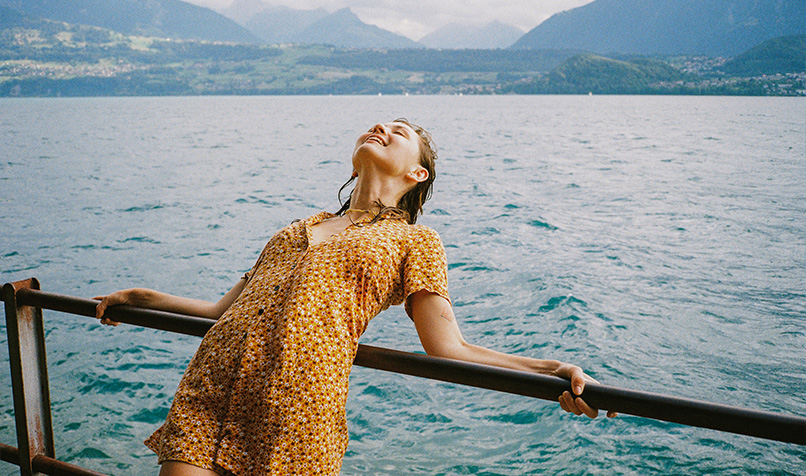 Person leaning back against a railing beside a lake, with mountains visible in the distance.