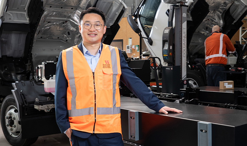 Person in a high-visibility vest standing inside a truck workshop beside a vehicle undergoing maintenance.