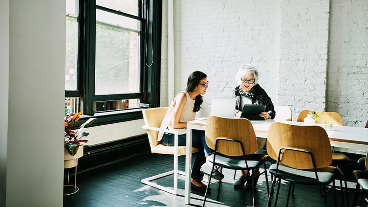 Two women desk talking looking tablet