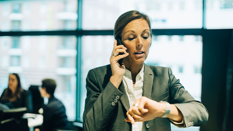 Busy businesswoman talking on mobile phone while checking time on watch at office