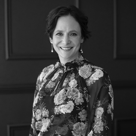 Black-and-white portrait of a person standing in front of dark panelled walls. The person is wearing a high-neck floral blouse with a detailed rose pattern and drop earrings. The image is framed from the waist up, with soft lighting highlighting the patterned clothing against the plain background.