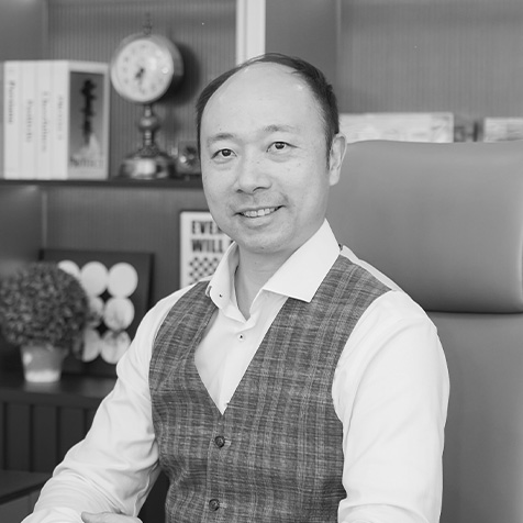 Black-and-white portrait of a person seated at a desk in an office. The person is wearing a white shirt and a checked waistcoat, facing the camera. Shelving in the background contains books, decorative objects and a small clock, creating a professional office setting.