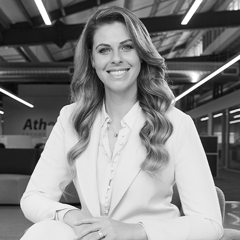Professional portrait of a person seated in a modern office, wearing a light-coloured blazer and looking towards the camera, with contemporary lighting and workspaces in the background.