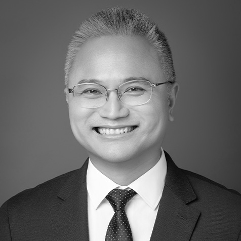 Black-and-white professional headshot of a person wearing glasses, a dark suit jacket, a white shirt and a patterned tie. The portrait is centred against a plain studio background, with the face and upper body clearly visible and evenly lit.