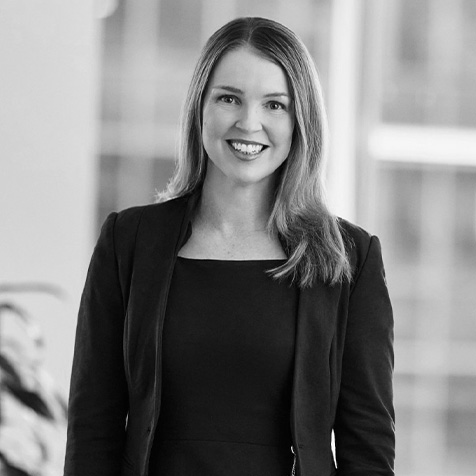 Black-and-white professional portrait of a person wearing a dark blazer, standing in an office environment with large windows in the background.