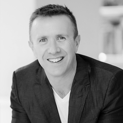 Man with short dark hair wearing a dark blazer leaning slightly forward smiling at camera
