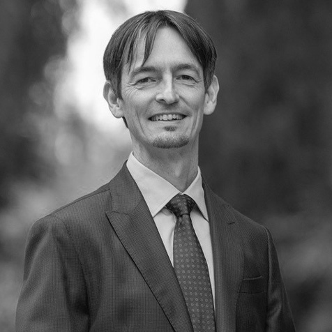 Black and white portrait of a person wearing a dark suit, light-coloured shirt and patterned tie, standing outdoors and facing the camera, with a softly blurred background of trees.