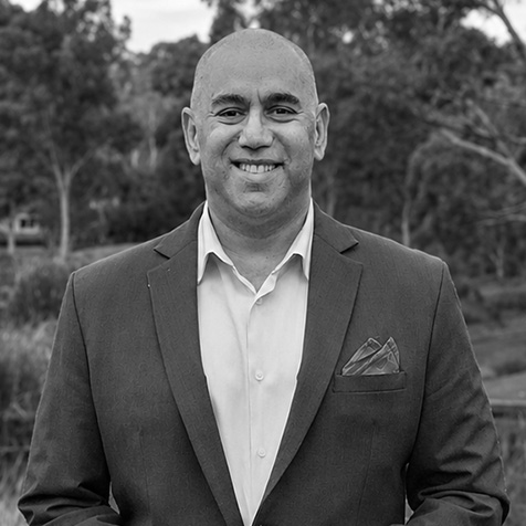 Head-and-shoulders portrait of a person wearing a suit jacket and collared shirt outdoors, with trees in the background.