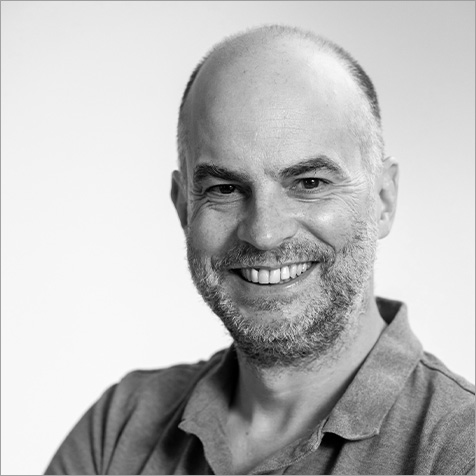 Professional head-and-shoulders portrait of a person against a light grey background. The person is wearing a blue polo shirt and is facing the camera with arms folded. The image is sharply focused with soft lighting, creating a friendly and approachable corporate profile photograph.