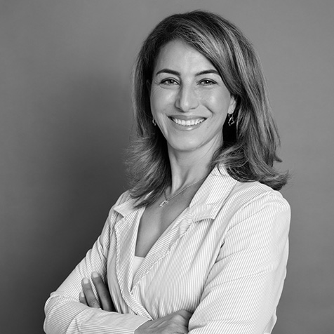 Professional studio portrait of a person standing with arms crossed against a plain grey background. The person is wearing a light blue striped wrap-style blazer over a white top, with small gold earrings and a delicate necklace. The image is well lit and framed from the waist up, creating a polished corporate headshot.