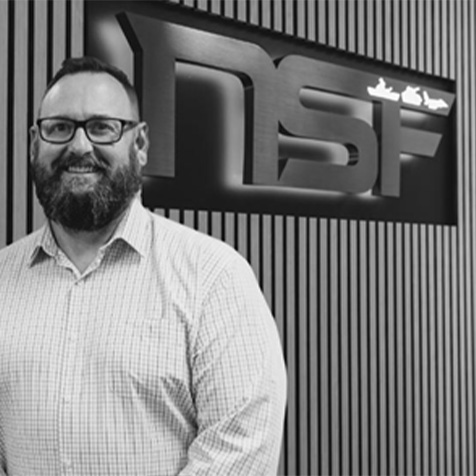 Black and white image of man in light coloured shirt with a beard and wearing glasses. Standing in front of a wall with NSF logo