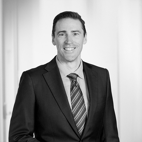 Professional headshot of a person wearing a dark business suit and striped tie, standing in an office setting.