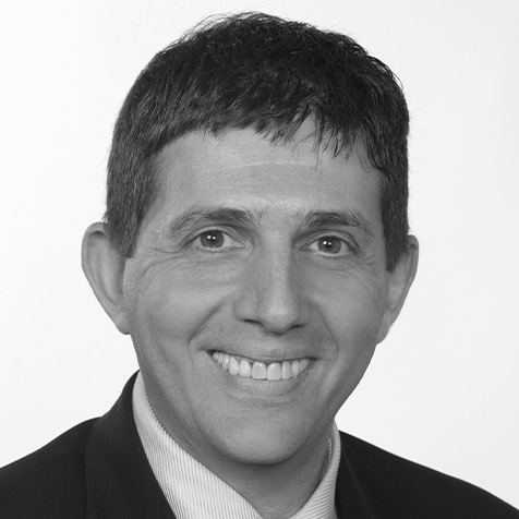 Professional headshot of a person wearing a dark suit jacket, light blue striped shirt and blue patterned tie, against a plain light grey background.