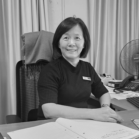 Black-and-white photograph of a person seated at an office desk, wearing a dark short-sleeved top, with a computer, telephone and desk fan visible in the workspace.
