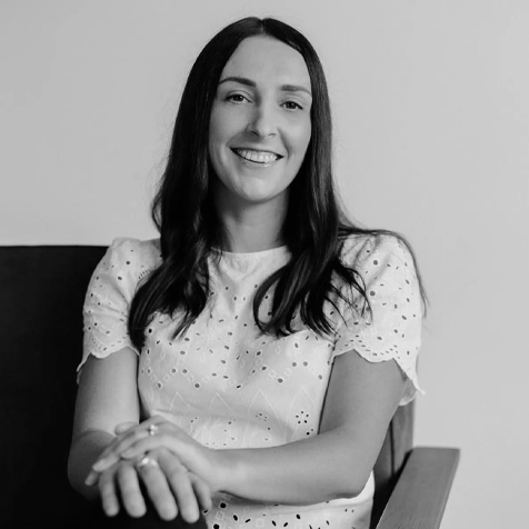 Black-and-white portrait of a person with long dark hair seated in a chair, wearing a white short-sleeved embroidered top, with hands folded in front.