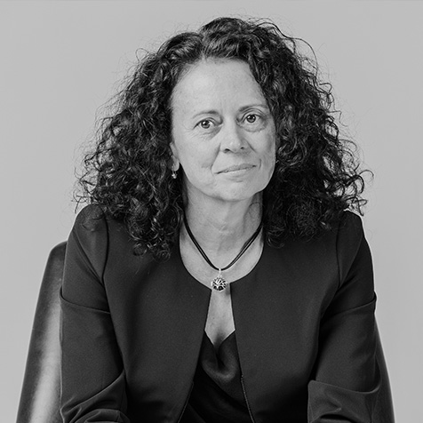 Black-and-white portrait of a person with curly hair seated in a chair, wearing a dark jacket and pendant necklace.