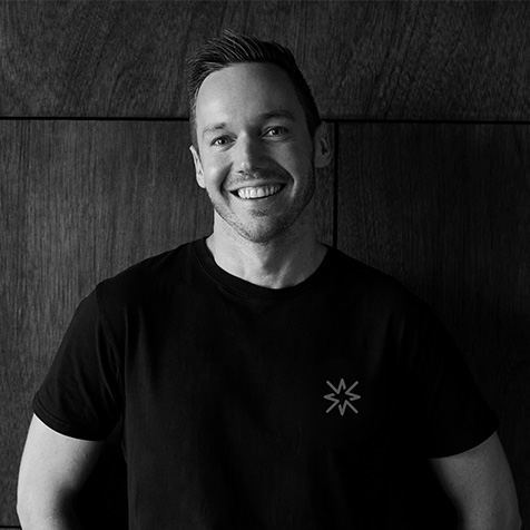 Professional black-and-white photograph of a person in a plain dark T-shirt, standing against a dark timber-panelled background, with the upper body visible and the face directed towards the camera.
