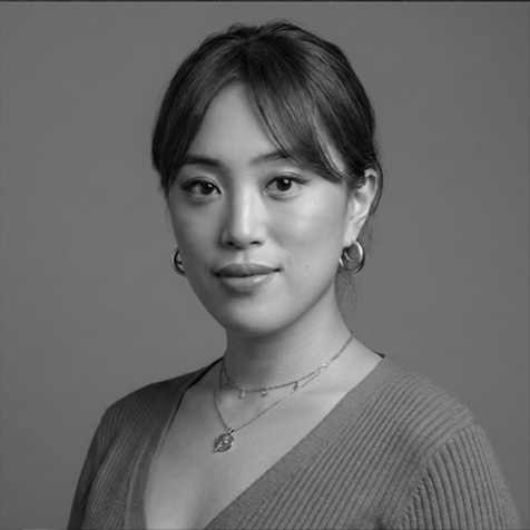 Black-and-white head-and-shoulders portrait of a person with dark hair tied back, wearing hoop earrings, layered necklaces and a ribbed V-neck top, facing the camera against a plain background.