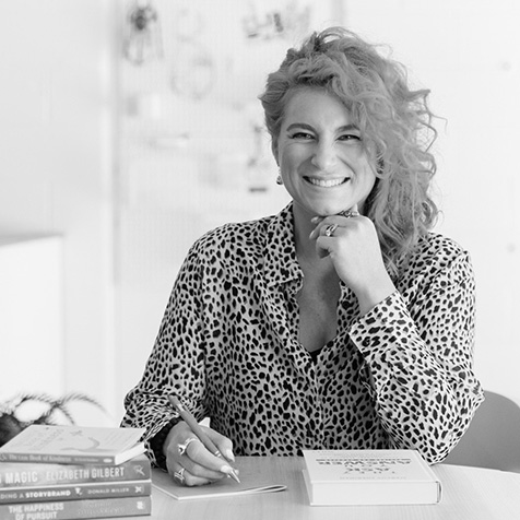Person seated at a desk writing in a notebook, with a stack of books in the foreground and a blurred office background. Black-and-white photograph.
