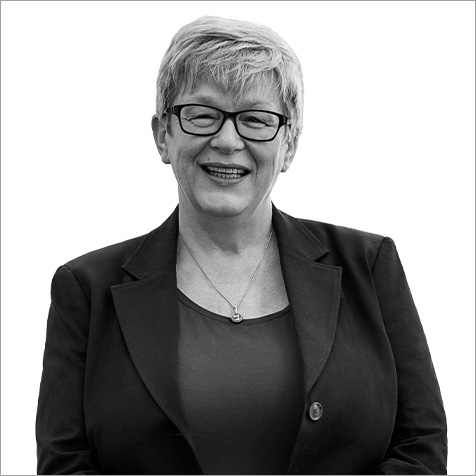 Black-and-white portrait of a person wearing glasses, a dark blazer, and a top, facing the camera against a plain light background, with short, neatly styled hair and a visible necklace.