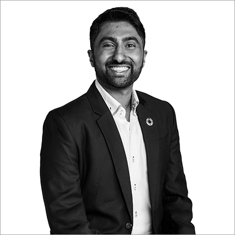 Black-and-white portrait of a person with short hair and a neatly trimmed beard, wearing a dark blazer over a light collared shirt. The person is seated at an angle and facing the camera against a plain light background, with a circular lapel pin visible on the blazer.