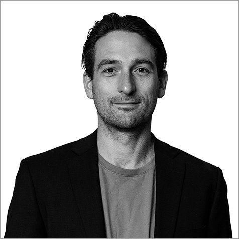 Black-and-white portrait of a person with short, neatly styled hair, wearing a dark blazer over a plain crew-neck shirt, facing forward against a plain light background.