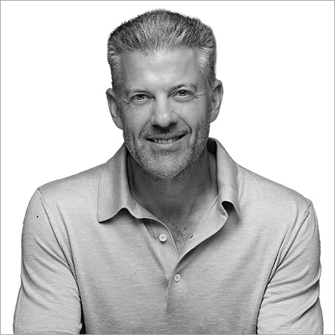Black-and-white portrait of a person with short, neatly styled hair and light facial stubble, wearing a light-colored collared polo shirt. The person faces the camera against a plain light background with arms resting forward.