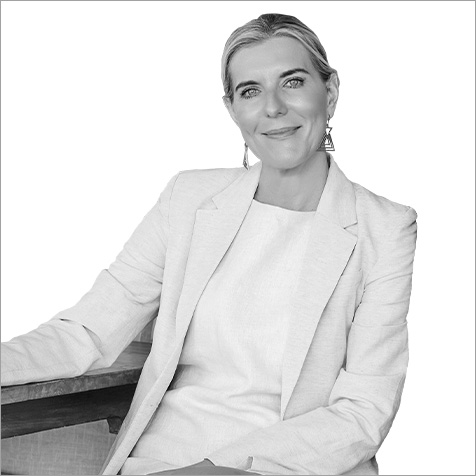 Studio portrait of a person wearing a light-coloured blazer and geometric earrings, seated beside a wooden table against a plain background.