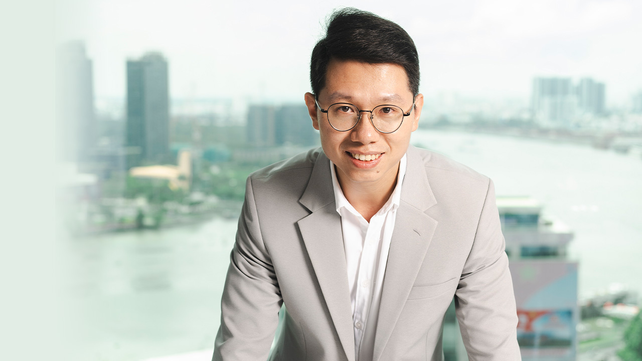 Person wearing glasses and a light-coloured suit jacket, photographed indoors with a blurred city and river view visible through windows in the background.