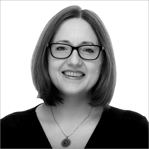 Black-and-white headshot of a person with shoulder-length hair wearing rectangular glasses, a dark V-neck top and a pendant necklace. The portrait is cropped from the chest upwards against a plain light background, with the subject facing the camera. The simple composition and neutral backdrop keep the focus on the person's face and professional appearance.