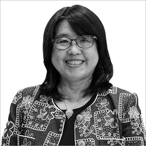 Black-and-white professional headshot of a person wearing glasses, a patterned jacket with decorative embroidered-style detailing, a dark top and a pendant necklace. The portrait is cropped from the chest upwards against a plain light background, with the subject facing the camera. The intricate floral and geometric patterns on the jacket contrast with the simple backdrop, creating a formal and professional profile image.