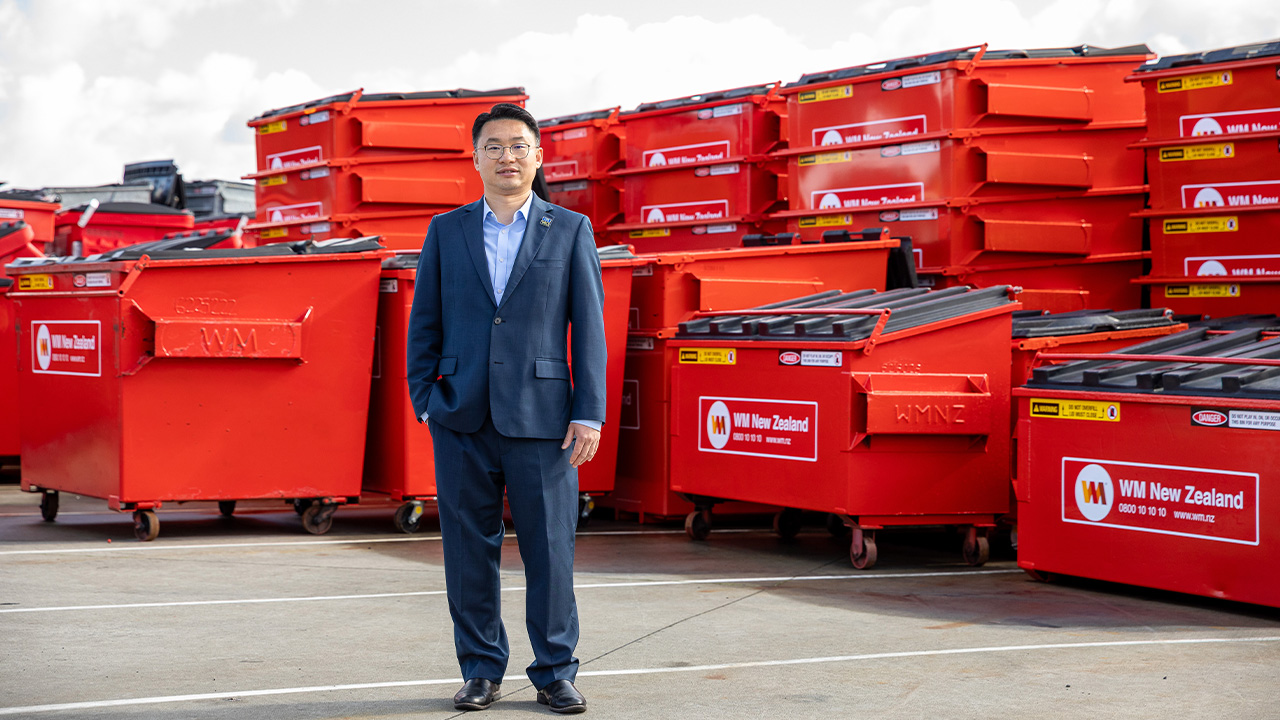 Person standing in a waste management depot among rows of red WM New Zealand skip bins.